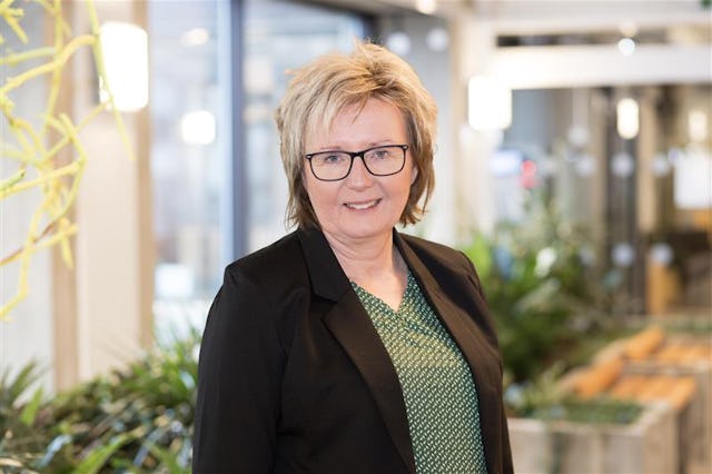 Helena Rödén med kort blont hår och glasögon står i en inomhusmiljö med växter och dagsljus. Hon har på sig en svart kavaj och en grönmönstrad blus.
