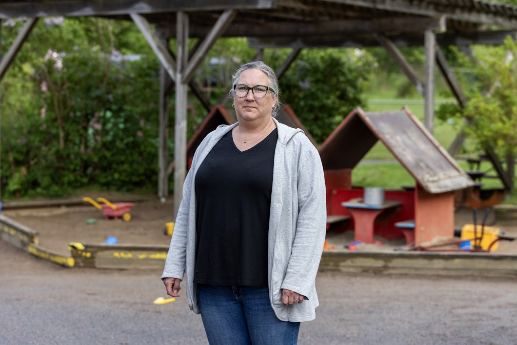 Malin Kungshammar med glasögon, svart tröja och grå huvtröja står framför en lekplats utomhus med små lekhus och utspridda leksaker.