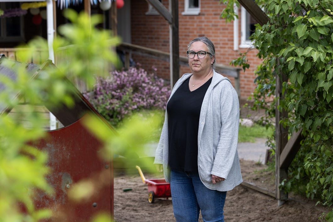 Malin Kungshammar med glasögon, grå kofta och jeans står utomhus i en trädgård med en röd tegelbyggnad och växter i bakgrunden.