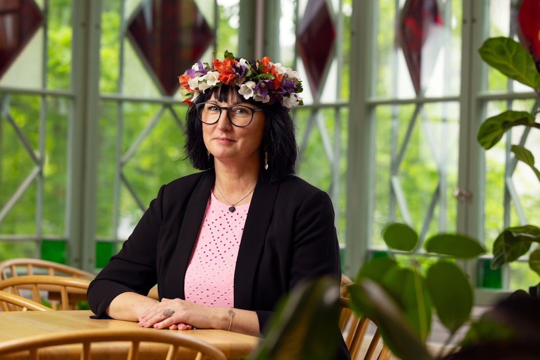 Anna Brandenberg med glasögon, mörkt hår och en blomkrans sitter vid ett bord i ett rum med stora fönster och växter.