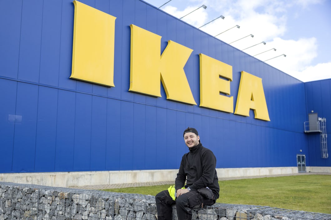 En person i svarta kläder sitter på en stenmur framför en stor blå IKEA-butik med tydliga gula skyltar.