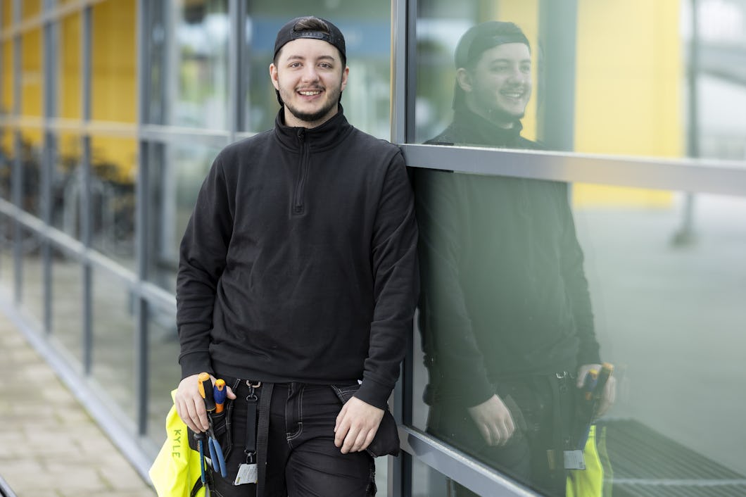 En person med svarta kläder och keps står vid en glasvägg, ler, och håller i en säkerhetsväst samt verktyg. Personens spegelbild syns i glaset.