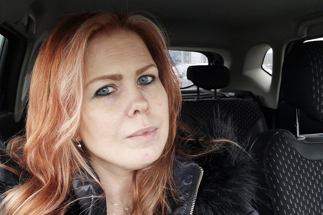 En kvinna med långt rödaktigt hår och ljus hud sitter i förarsätet på en bil och tittar in i kameran med ett neutralt ansiktsuttryck.