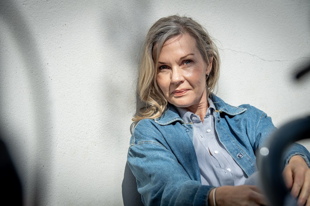 Pia Johansson med blont hår sitter mot en vit vägg. Hon har på sig en jeansjacka och en ljusblå skjorta och tittar in i kameran i dagsljus.