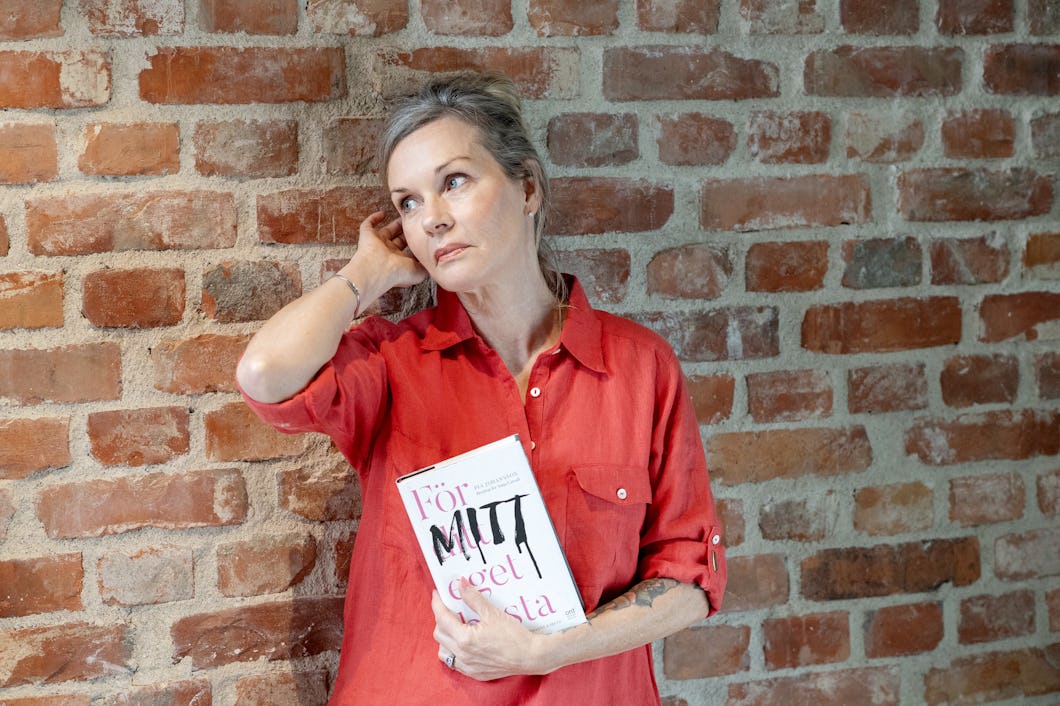 Pia Johansson med en röd skjorta står mot en tegelvägg, håller i en bok med titeln ”För ditt eget bästa” och vänder blicken åt sidan.