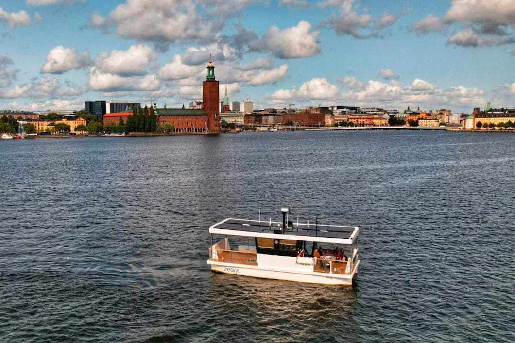 En båt över vattnet. I bakgrunden syns en stad med ett tydligt torn. Himlen har både moln och klara partier.