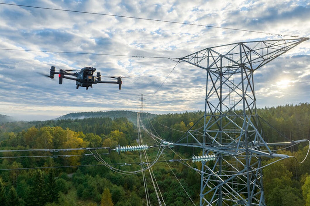 En drönare flyger nära kraftledningar och en transmission torn för att inspektera elinfrastruktur ovanför ett skogsområde under en delvis molnig himmel.