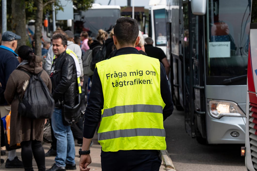 En person med en väst med texten "Fråga mig om tågtrafiken!" står nära ett område där det finns många människor och bussar.