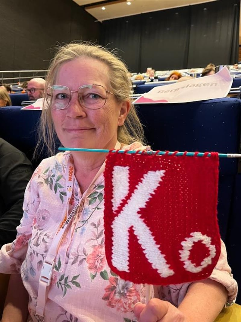 Lena Eldstål med glasögon sitter i en aula och håller upp en rödvit stickning med Kommunals logga.