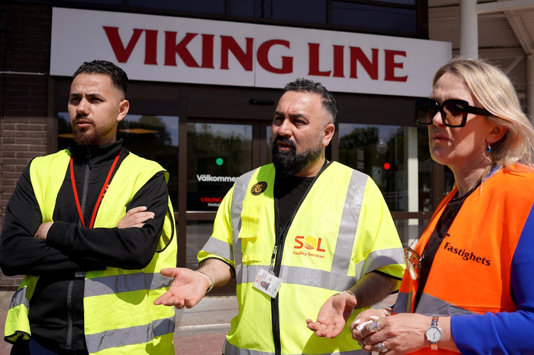 Tre personer med säkerhetsvästar och brickor står utanför en Viking Line-terminal och pratar med varandra.