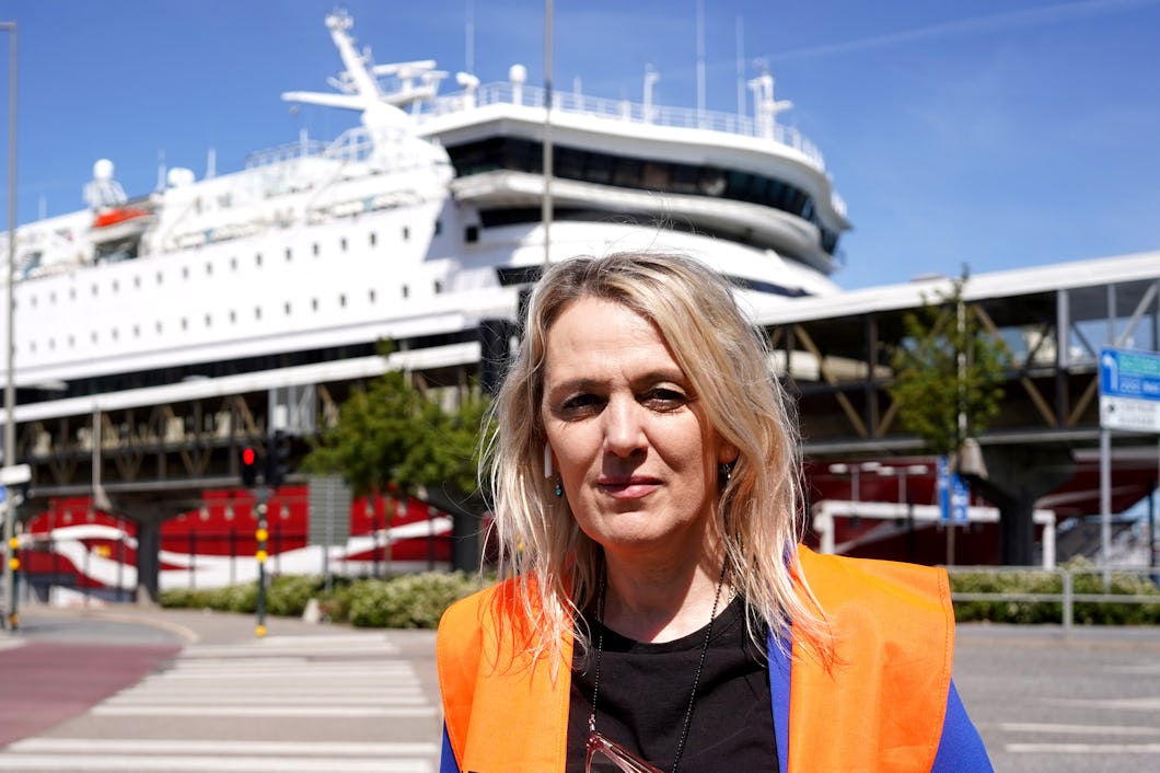 En kvinna med en orange säkerhetsväst står utomhus. I bakgrunden ligger ett vitt kryssningsfartyg vid kaj.