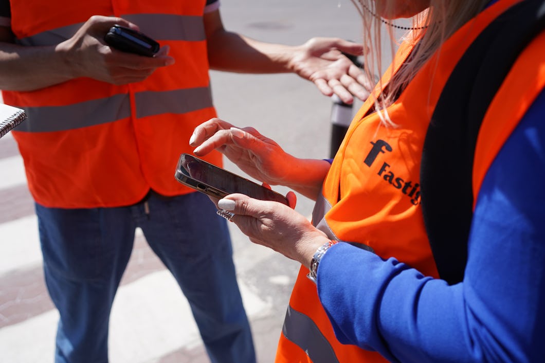 Två personer med orange säkerhetsvästar står utomhus; en skriver på en mobiltelefon medan den andra håller en telefon och gestikulerar med ena handen.