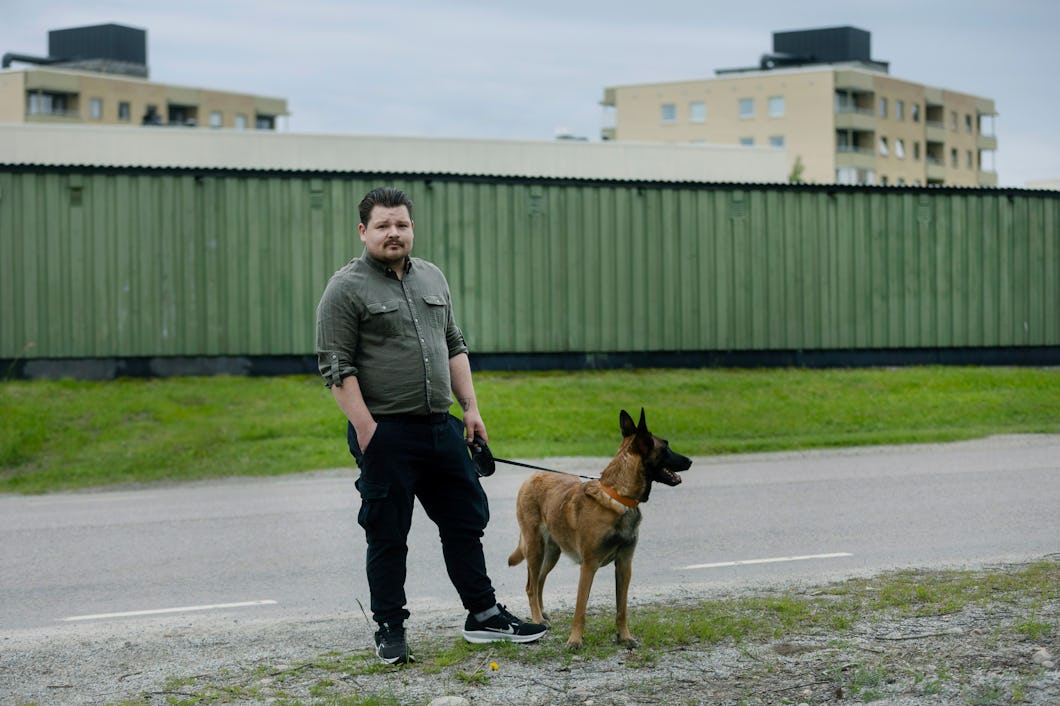 En man står vid vägkanten och håller i en koppel. Bredvid honom finns en brun hund. I bakgrunden syns hyreshus och ett grönt staket av metall.
