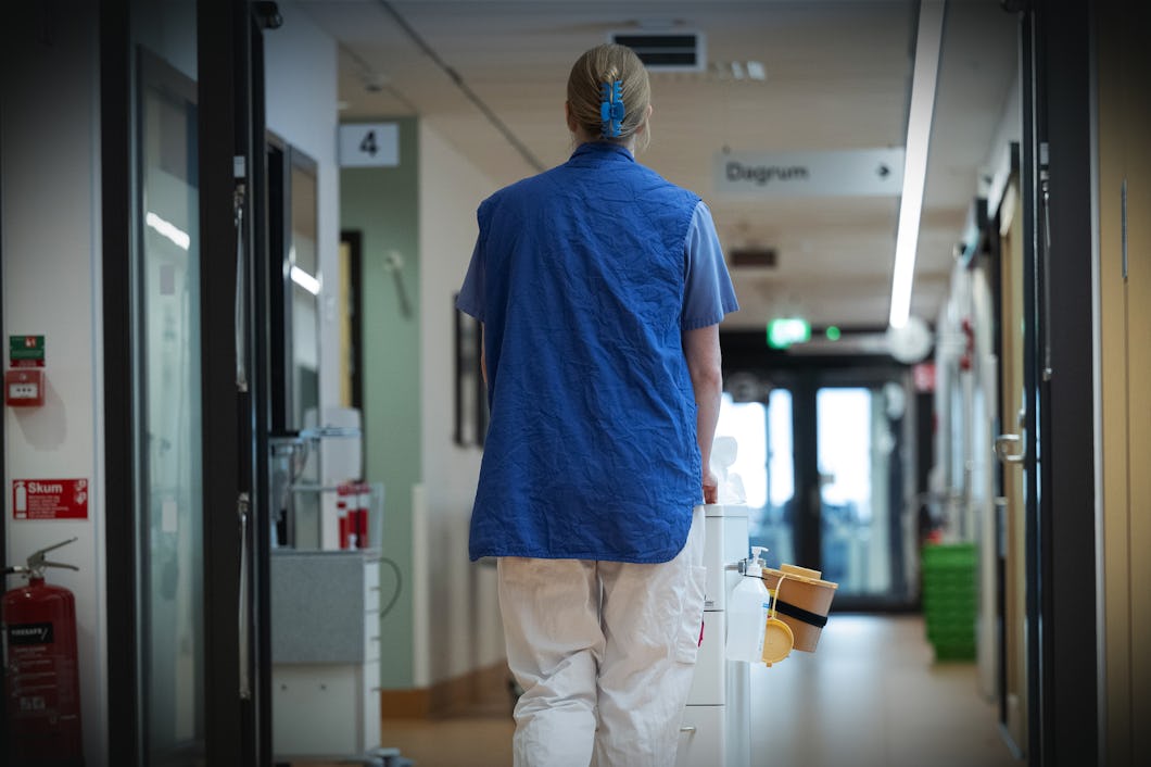 En undersköterska går bort från kameran i en sjukhuskorridor med medicinsk utrustning.