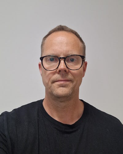 En man med kort hår och glasögon har på sig en svart tröja. Han står mot en vit bakgrund och tittar rakt in i kameran.
