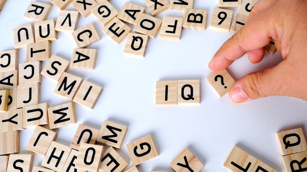 En hand placerar träbokstavsbrickor för att bilda "IQ?" bland utspridda bokstavsbrickor på en vit yta.