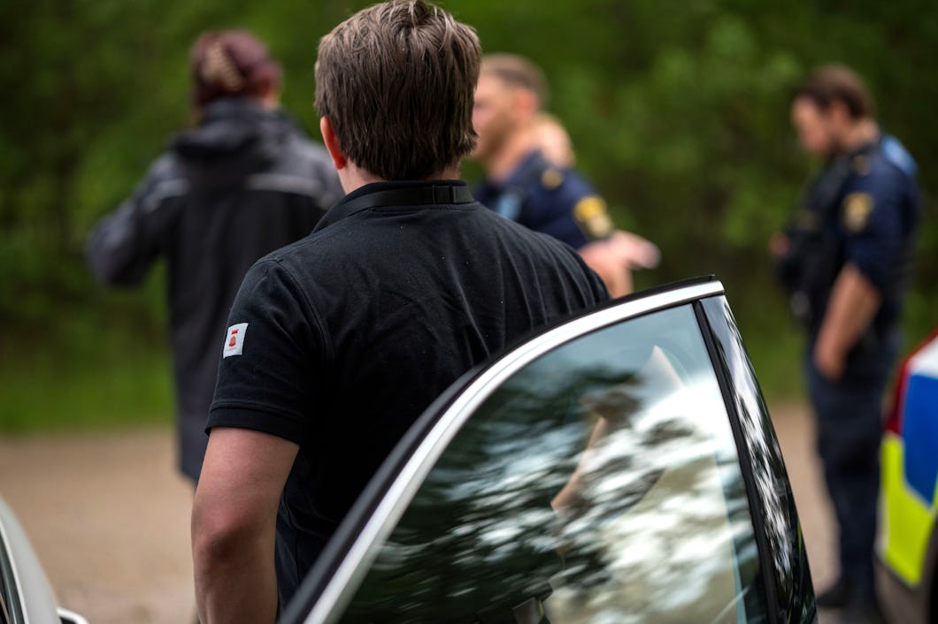 En person står vid en öppen bildörr medan flera personer, däribland två i polisuniform, samtalar i bakgrunden utomhus.