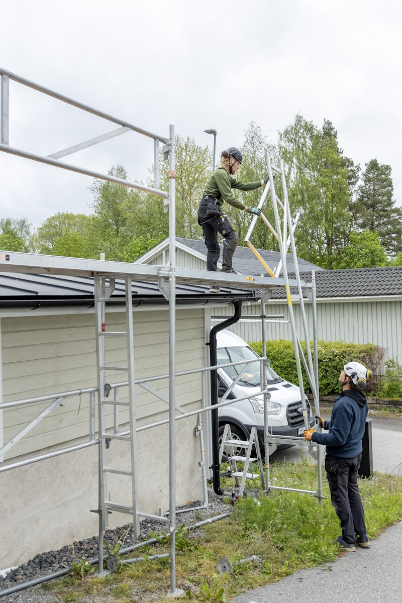 Två arbetare med hjälmar monterar metallställning vid ett hus. En person står på ställningen och den andra hjälper till från marken.