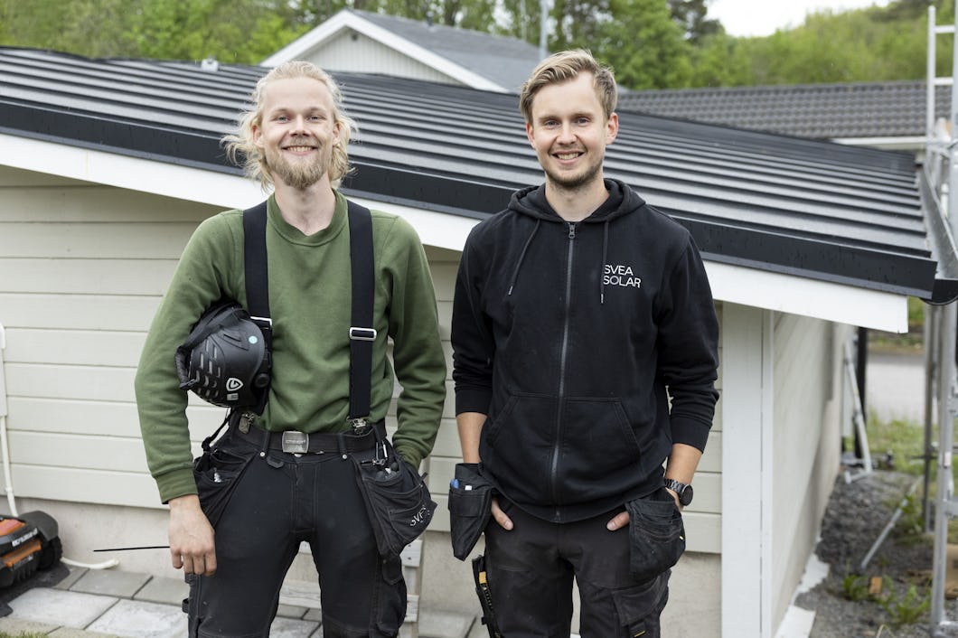 Två män i arbetskläder står framför en ljus byggnad med mörkt tak. Den ena mannen har på sig en grön tröja och ett verktygsbälte, den andra bär en svart huvtröja med texten "Svea Solar".