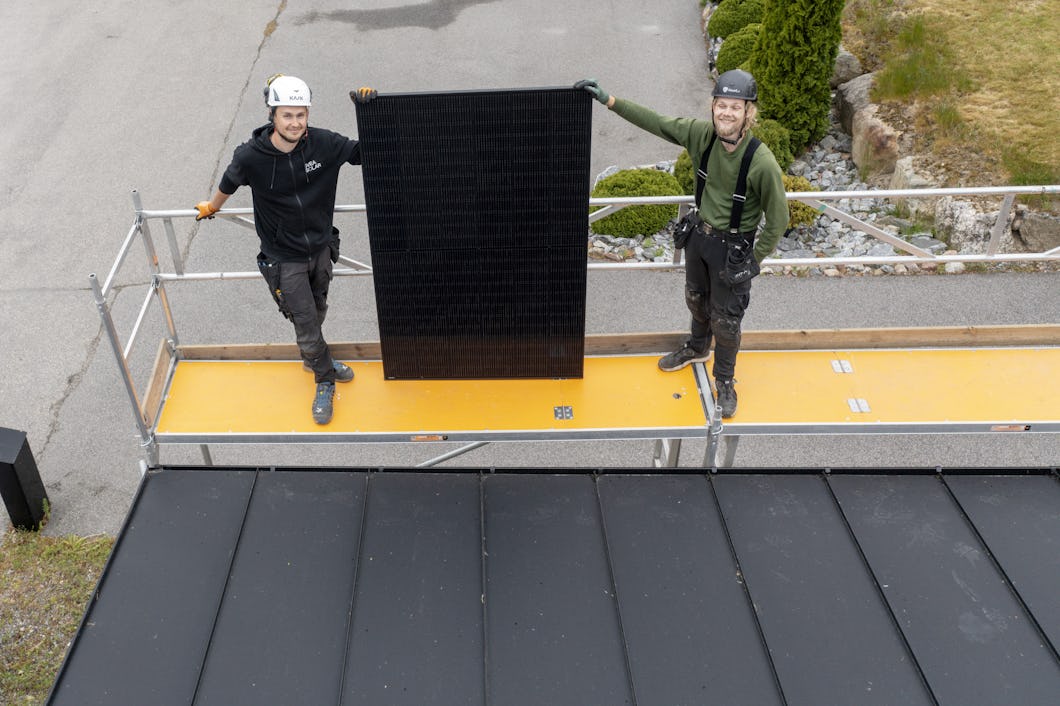 Två arbetare på en byggnadsställning håller upp en solpanel ovanför ett som delvis är täckt med monterade solpaneler.
