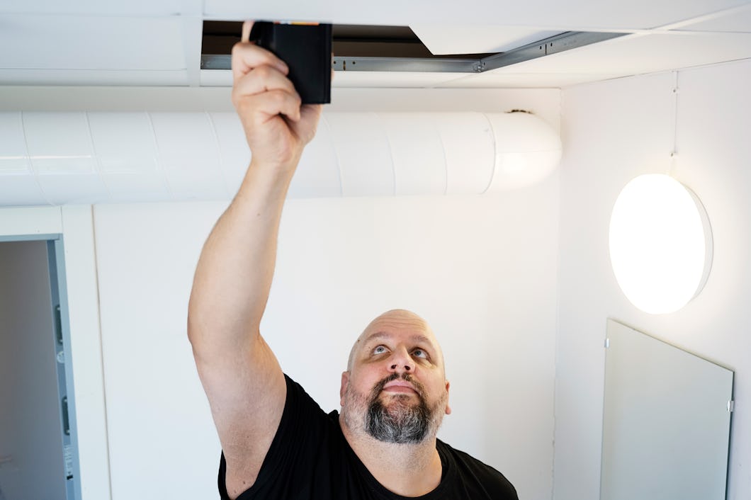 En man sträcker sig upp för att installera eller undersöka en apparat i ett öppet takpanel i ett rum med vita väggar och en rund vägglampa.