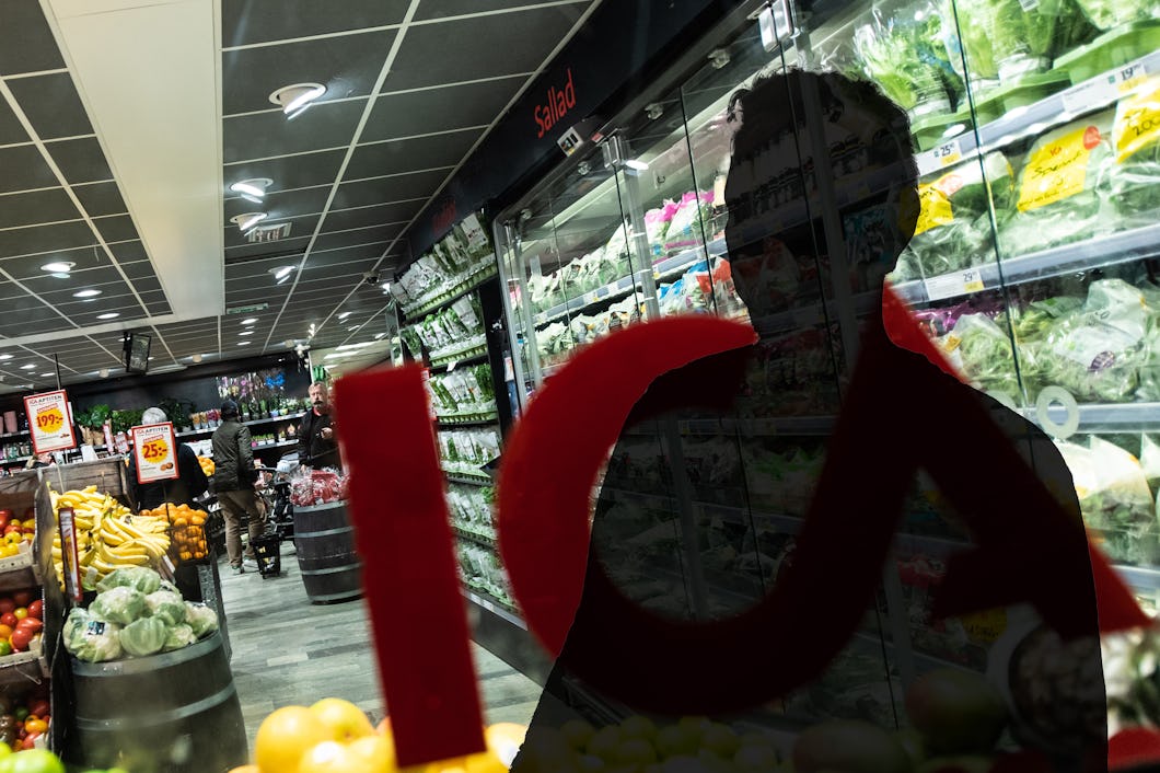 En frukt- och grönsaksavdelning i en mataffär syns genom ett fönster. På fönstret finns en del av en röd logotyp och en personsilhuett.