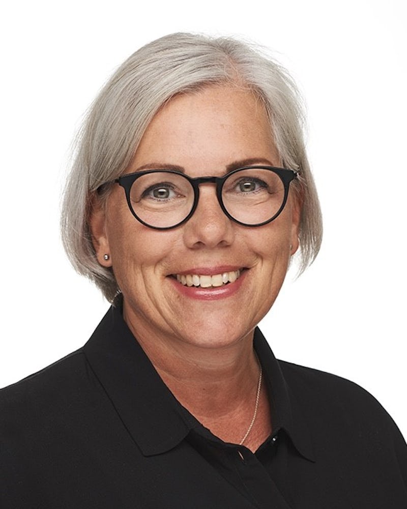 En kvinna med kort grått hår har runda svarta glasögon och en svart skjorta med krage. Hon ler mot kameran framför en vit bakgrund.