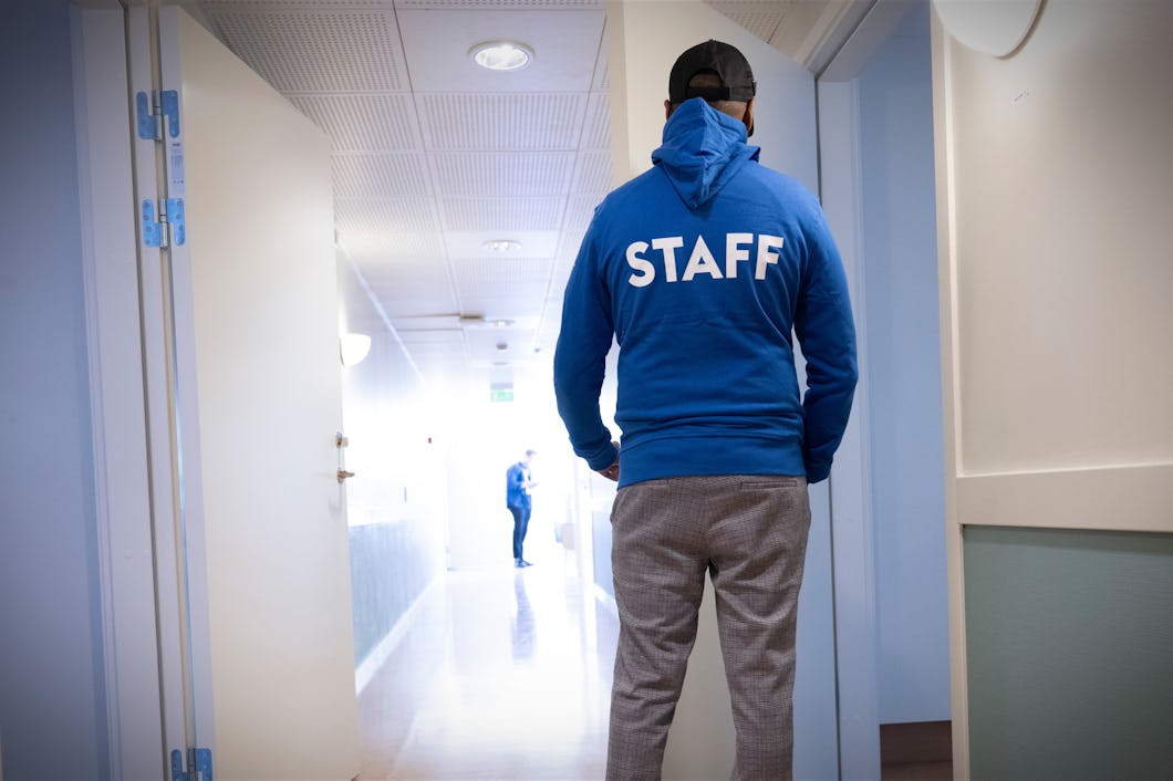 En person med en blå huvtröja med texten "STAFF" och grå byxor står i en korridor. En annan person syns längre bort i korridoren.