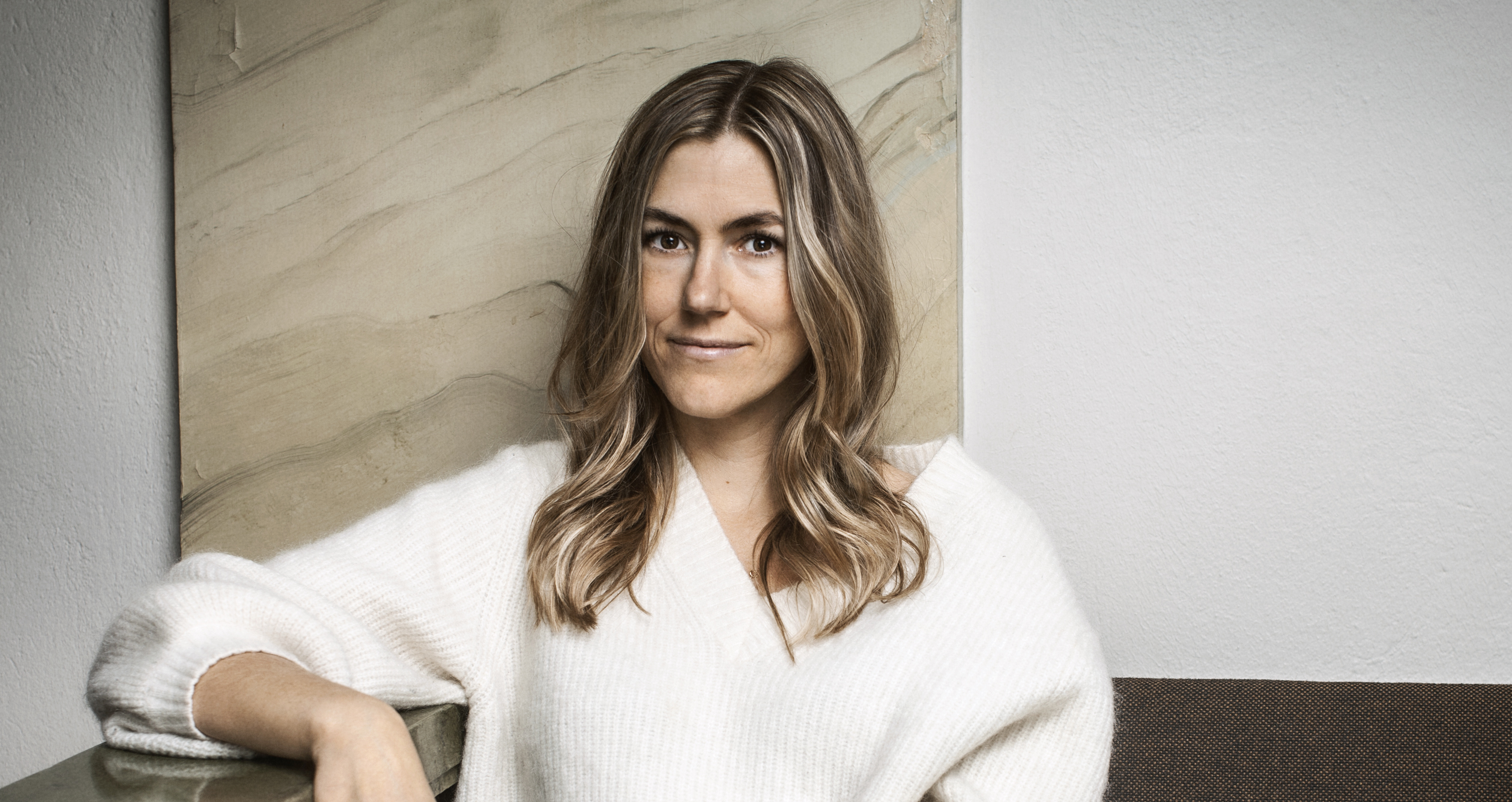 En kvinna med långt ljusbrunt hår och vit tröja sitter på en soffa framför en ljus vägg och abstrakt konst.