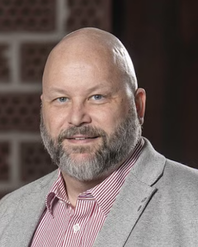 Patric Lundström med rakat huvud, skägg och mustasch har på sig en grå kavaj över en röd- och vitrandig skjorta. Han står framför en oskarp inomhusmiljö.