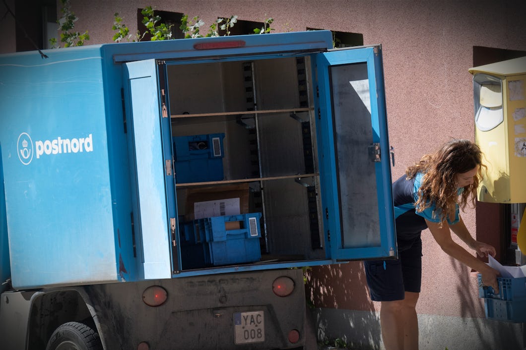 En brevbärare hämtar postbackar från ett PostNord-fordon med öppna dörrar vid en brevlåda på en gata i staden.