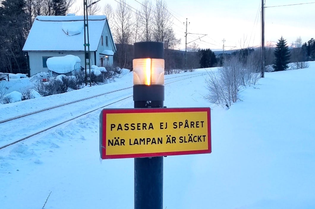 En tänd lampa sitter på en stolpe vid järnvägsspår med snö. En svensk skylt lyder: "Passera ej spåret när lampan är släckt.