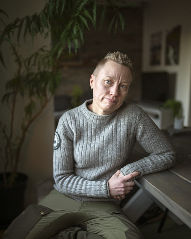 En person med kort hår har på sig en grå, ribbad tröja och gröna byxor. Personen sitter på en bänk inomhus och vilar ena armen på ett bord av trä.