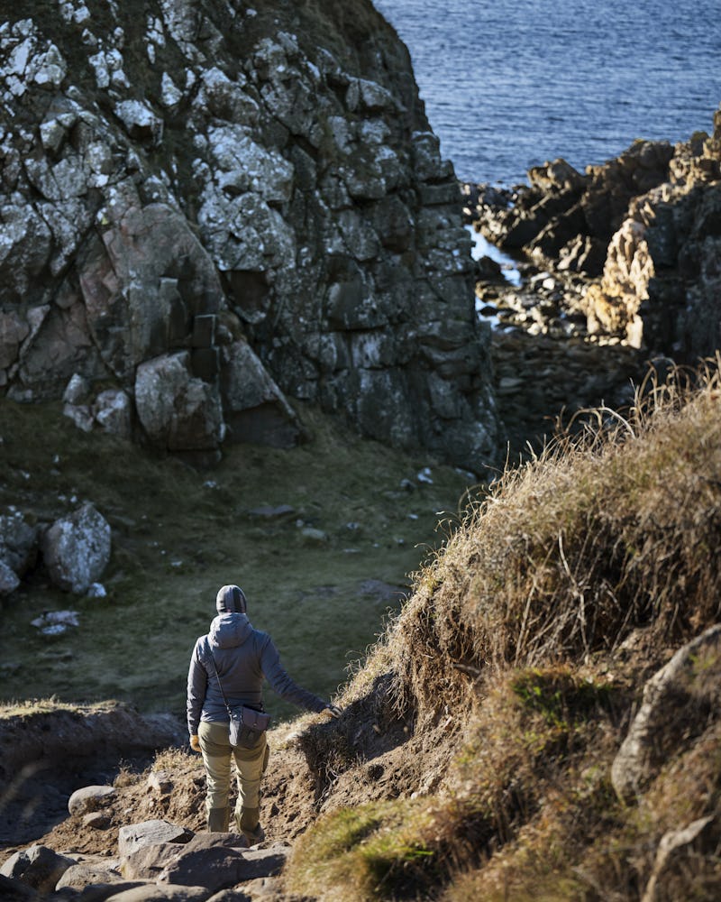 En person med jacka och ryggsäck går på en stenig stig mot en klippa vid kusten, med havet synligt i bakgrunden.