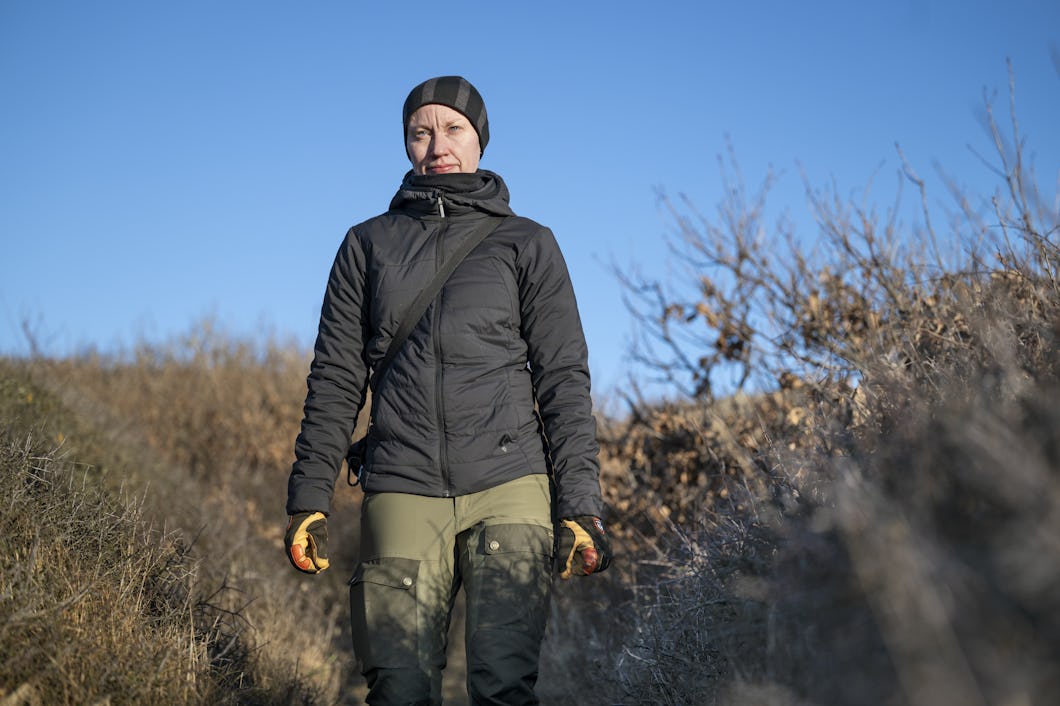 En person med svart jacka, gröna byxor, handskar och mössa står utomhus på en grusväg omgiven av torra buskar under en molnfri himmel.
