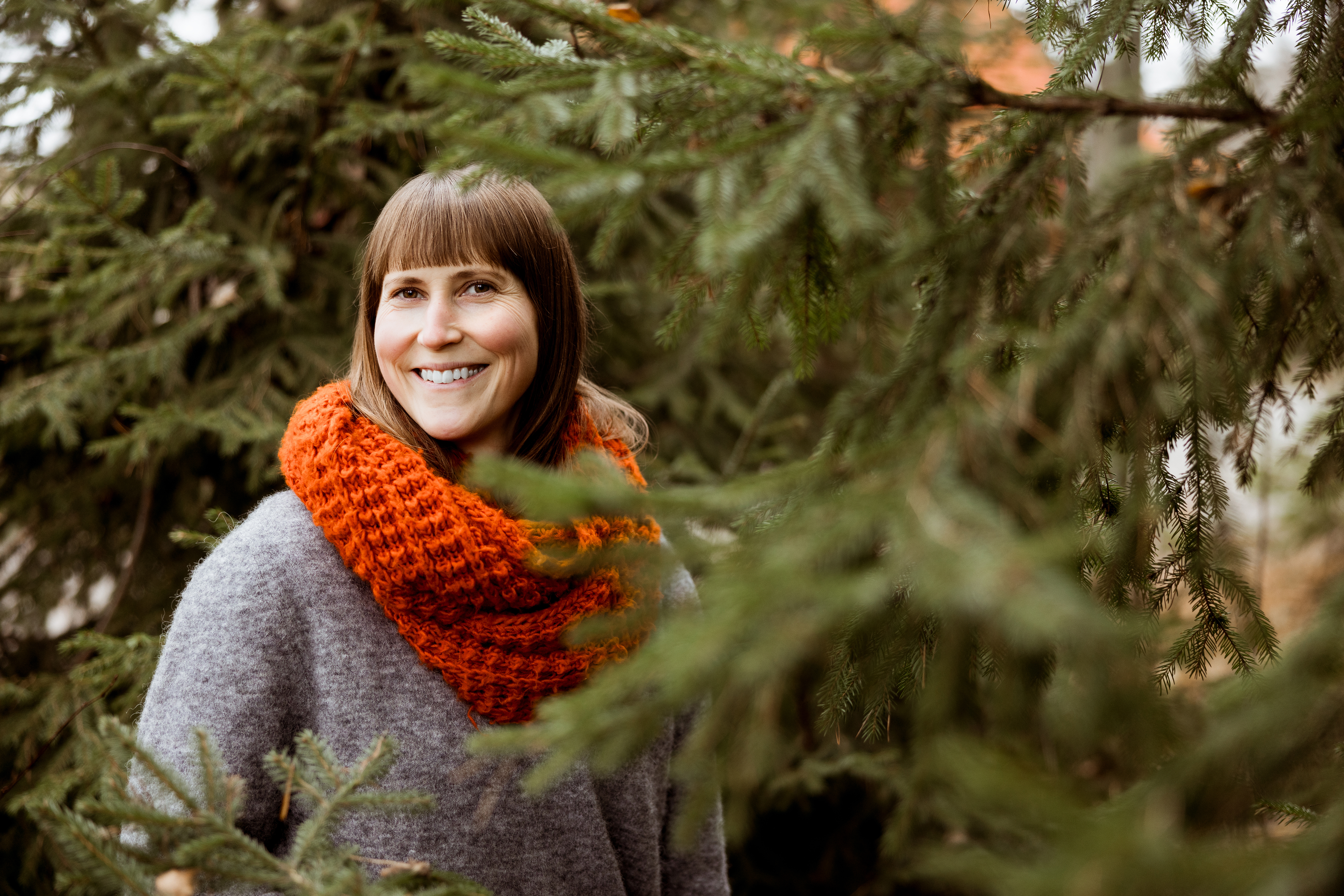 En kvinna med rakt brunt hår, klädd i en grå tröja och en orange halsduk, står bland barrträd och ler mot kameran.