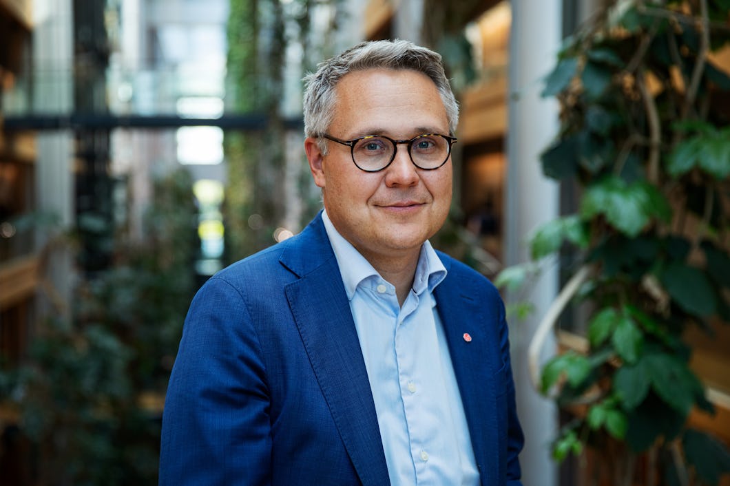 En man med glasögon och blå kavaj står inomhus i en byggnad med växter och glasväggar i bakgrunden.