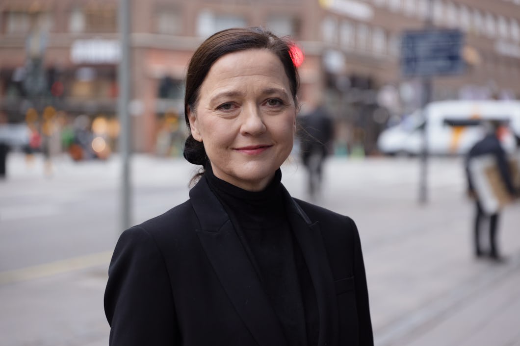 En kvinna i en svart kavaj står utomhus på en stadsgata och tittar in i kameran med ett neutralt ansiktsuttryck. I bakgrunden syns byggnader och människor, men de är inte i fokus.