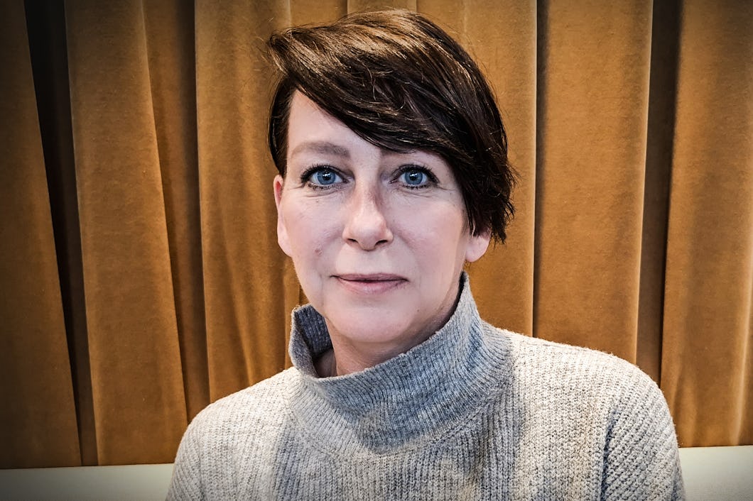 En person med kort brunt hår och blå ögon sitter framför guldfärgade gardiner och har på sig en grå polotröja.