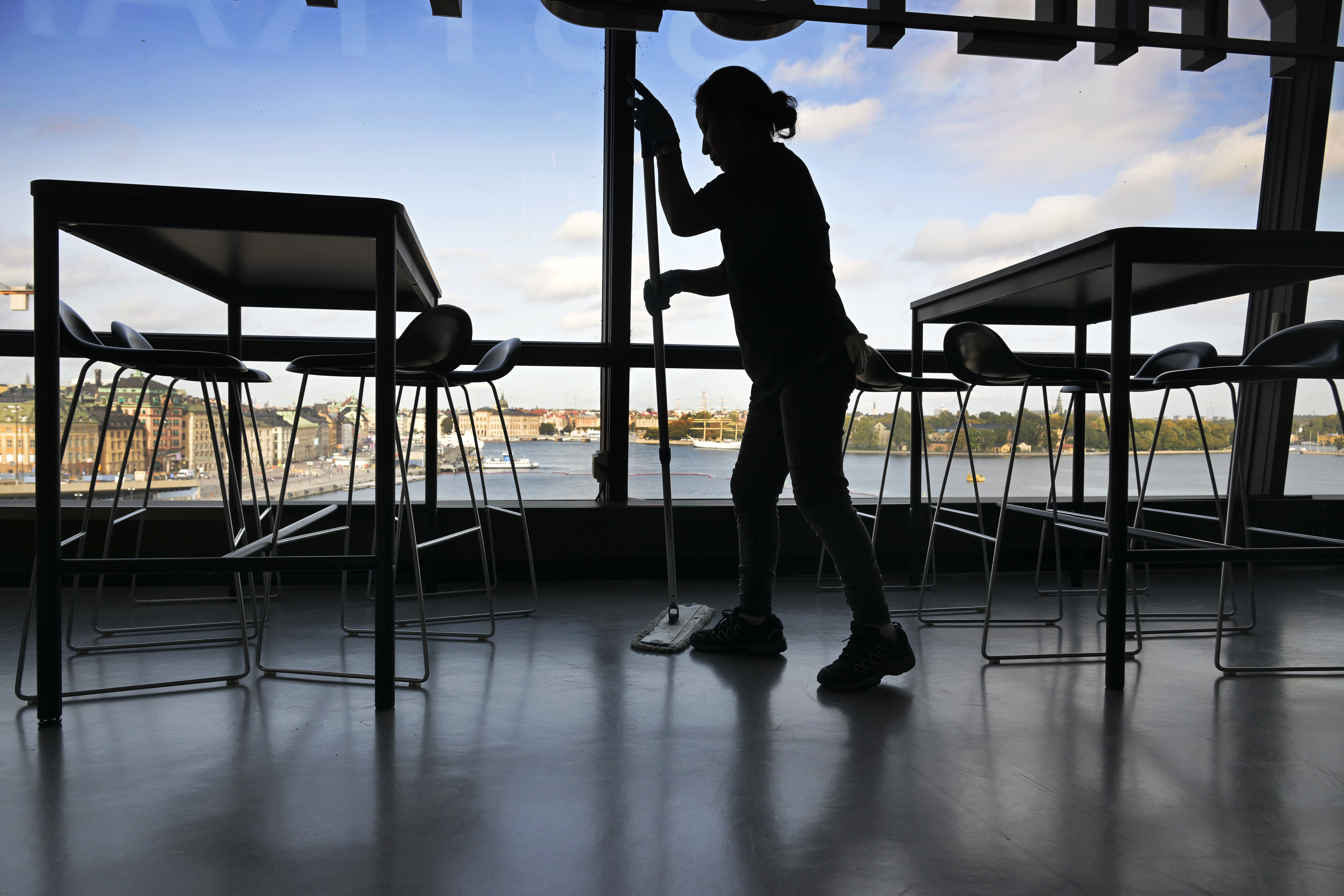 En person moppar golvet i en cafeteria med stora fönster som vetter mot staden och vattnet.