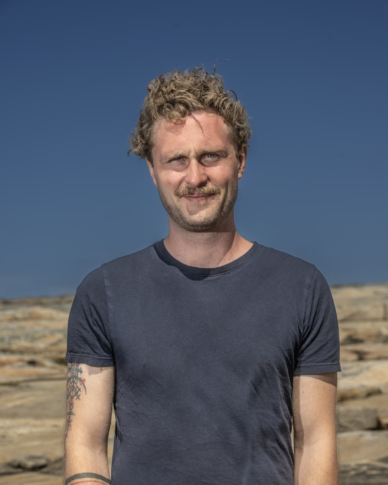 Brian Mortensen med lockigt hår och tatueringar på vänster arm står utomhus framför klippig terräng under en klar himmel.