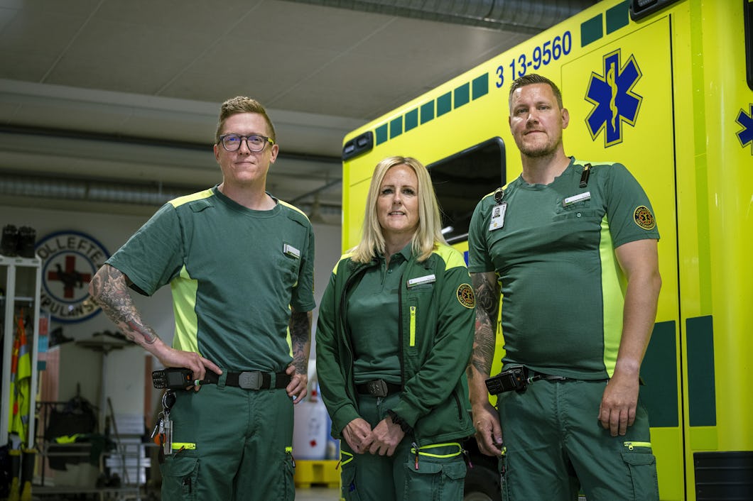 Linus Vestberg, Ulrika Maistedt och Tomas Sandberg står i gröna ambulanssjukvårdarkläder framför en gul ambulans inne i ett garage.
