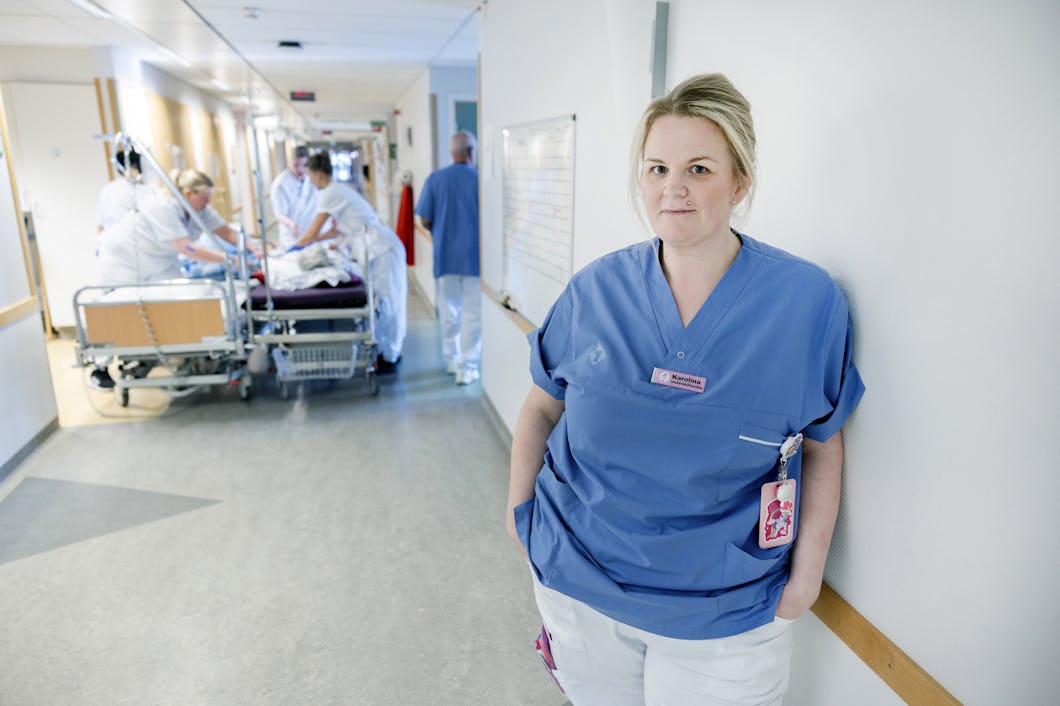 Karolina Persson i blå arbetskläder står i en sjukhuskorridor, medan medicinsk personal i bakgrunden tar hand om en patient på en bår.