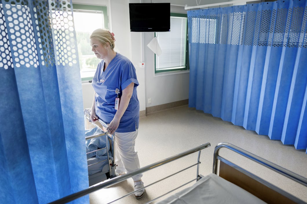 En undersköterska i arbetskläder står vid en sjukhussäng i ett patiententrum och tittar ut genom ett fönster med delvis fördragna gardiner.