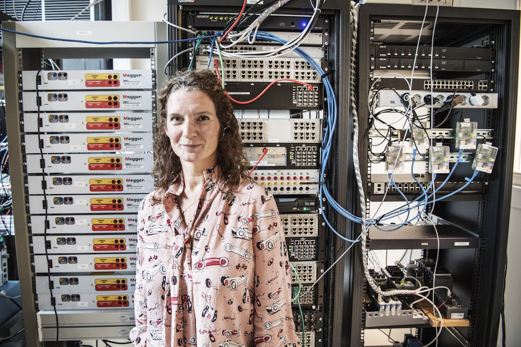 En kvinna står framför serverställ och nätverksutrustning, med kablar och elektroniska apparater synliga i en teknisk arbetsmiljö.