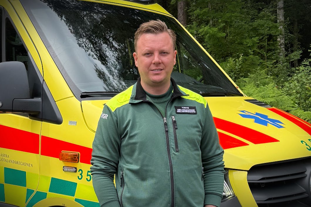 En man med en grön jacka står framför en ambulans som är parkerad utomhus nära träd och växter.
