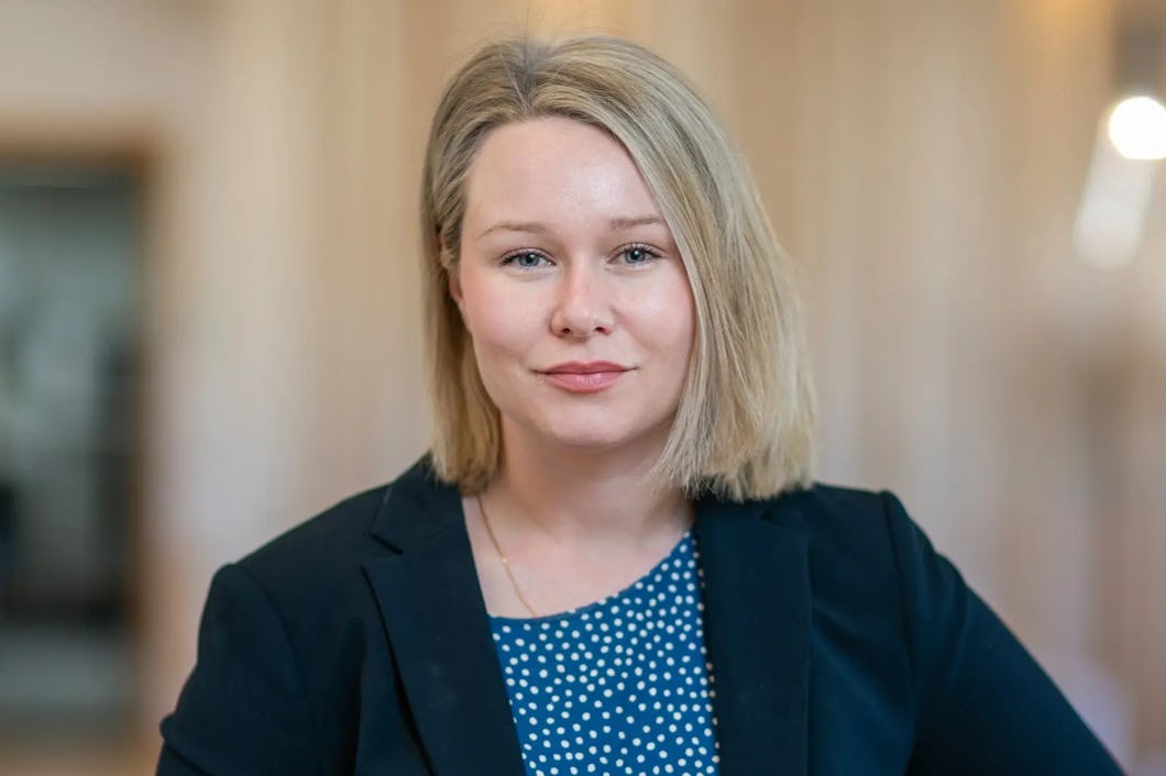 En kvinna med rakt blont hår, klädd i en mörkblå kavaj över en blus med blå prickar, står inomhus.