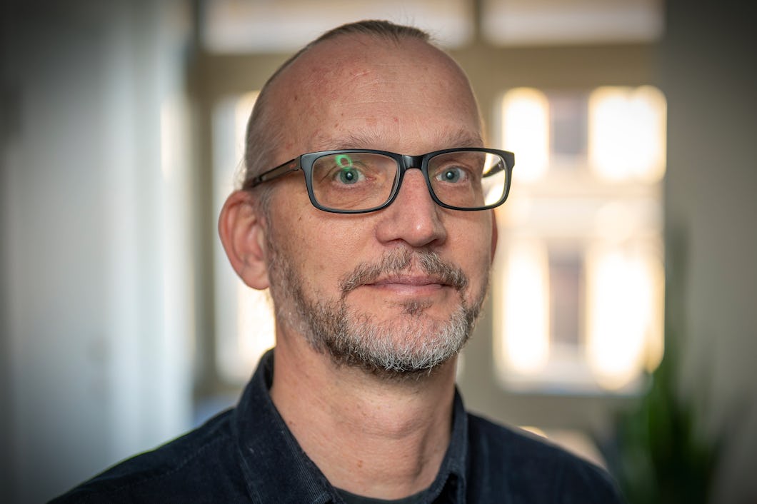 En man med glasögon, kort skägg och kort hår tittar mot kameran inomhus. I bakgrunden syns fönster.