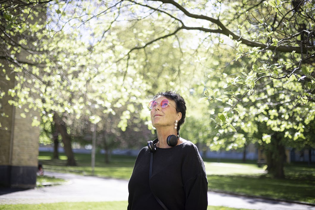 En person med rosa solglasögon och hörlurar står utomhus i en park och tittar upp mot träden.