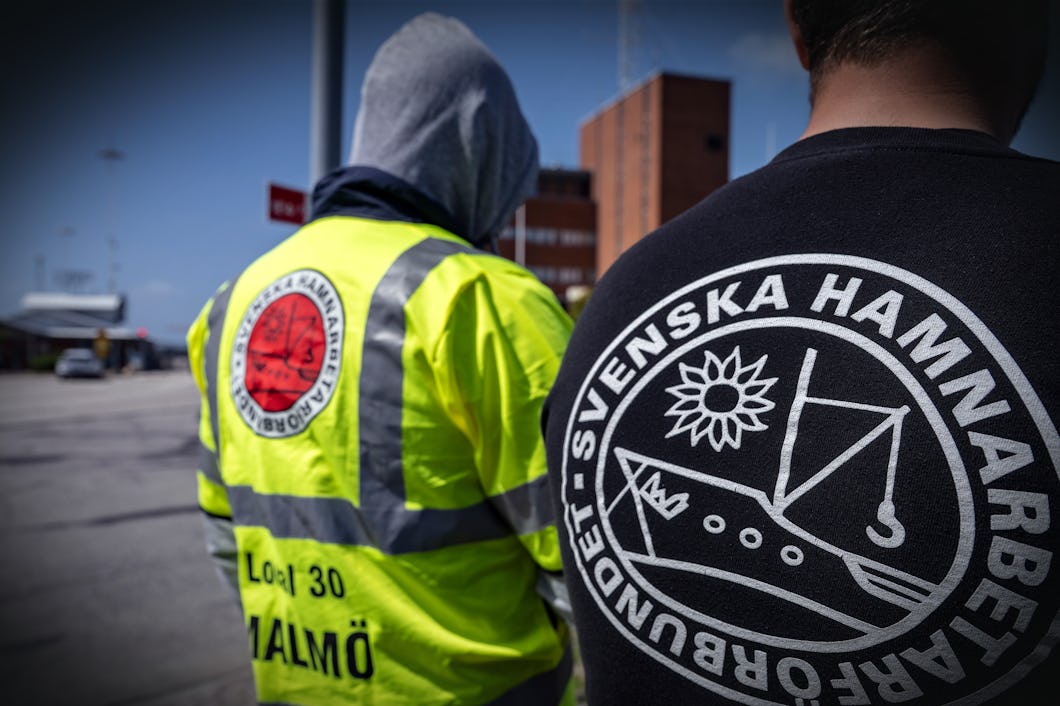 Två personer med jackor som har "Svenska Hamnarbetarförbundet" tryckt på sig står utomhus vid en byggnad i Malmö.